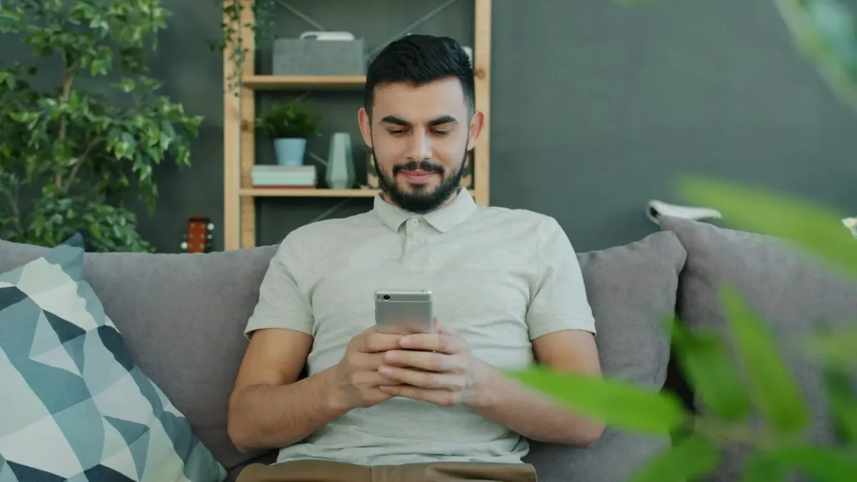 A man on a grey sofa at home, looking down at his phone, a plant in the foreground.