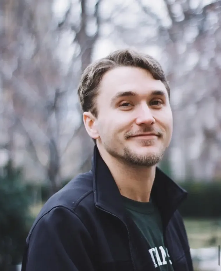 A young man in a dark zip-up jacket, smiling slightly, photographed outdoors among bare winter trees.