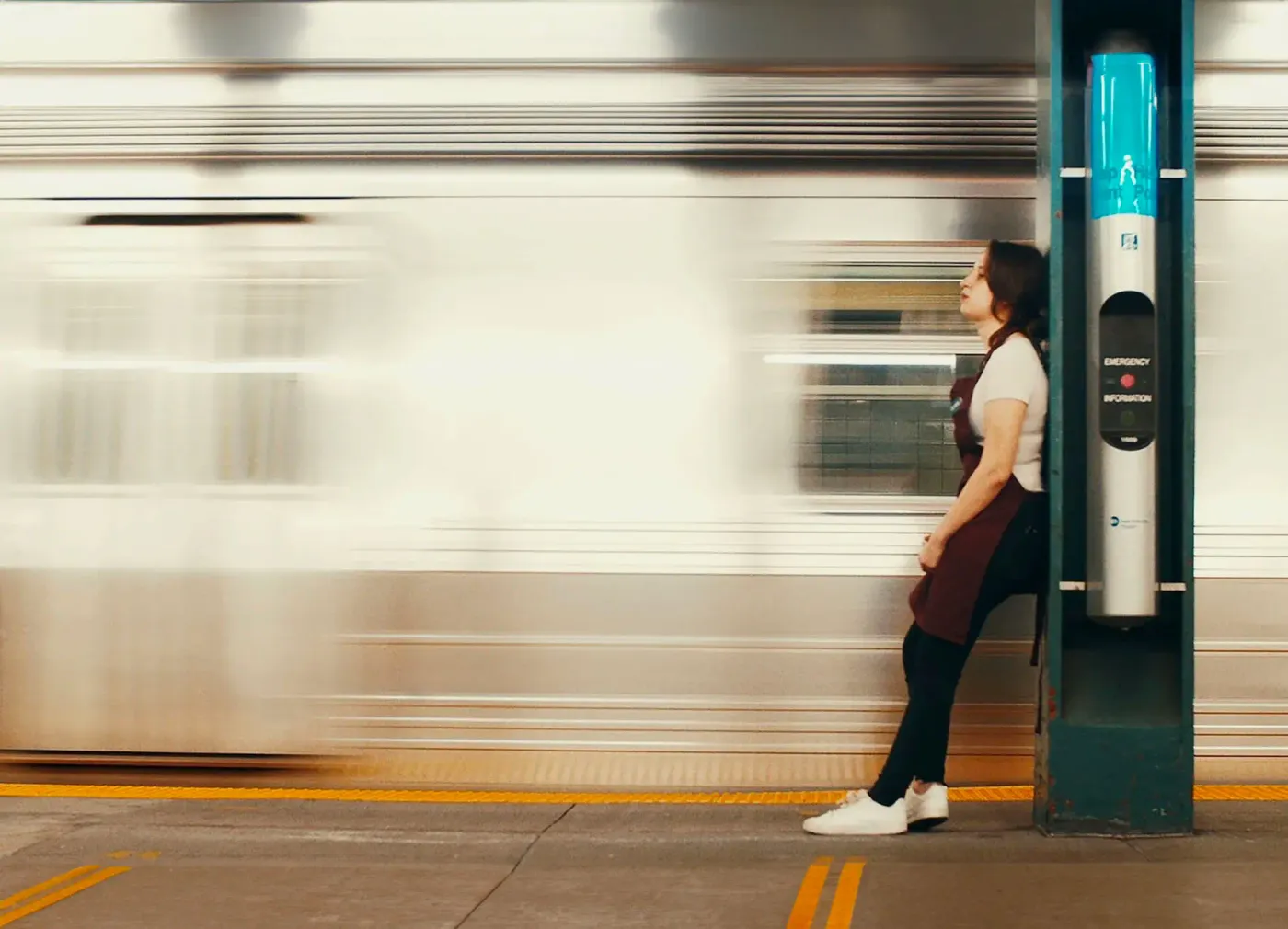 A woman in an apron leans against a post on a subway platform with her eyes closed while a train rushes past in a blur.