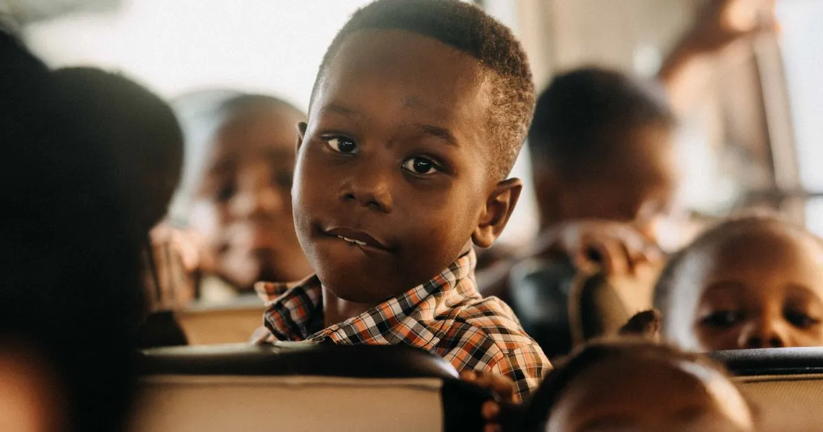 A boy in a plaid shirt looks back over the seat of a school bus, with other children around him.