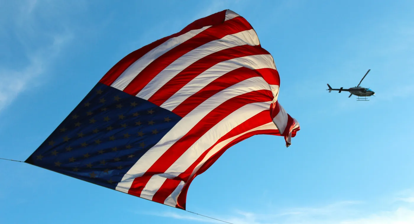 A large United States flag flying from a cable against a blue sky, with a helicopter in the distance.