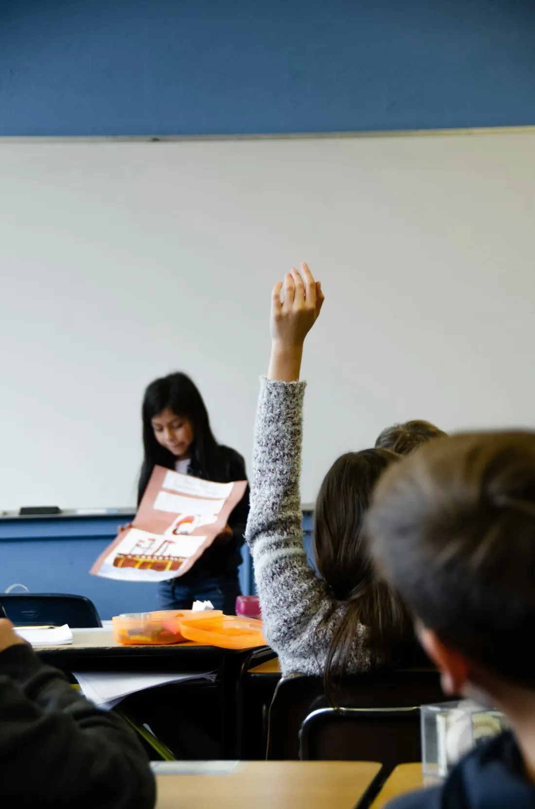 La mano de un estudiante levantada en un aula, vista desde atrás, mientras una compañera presenta al frente.