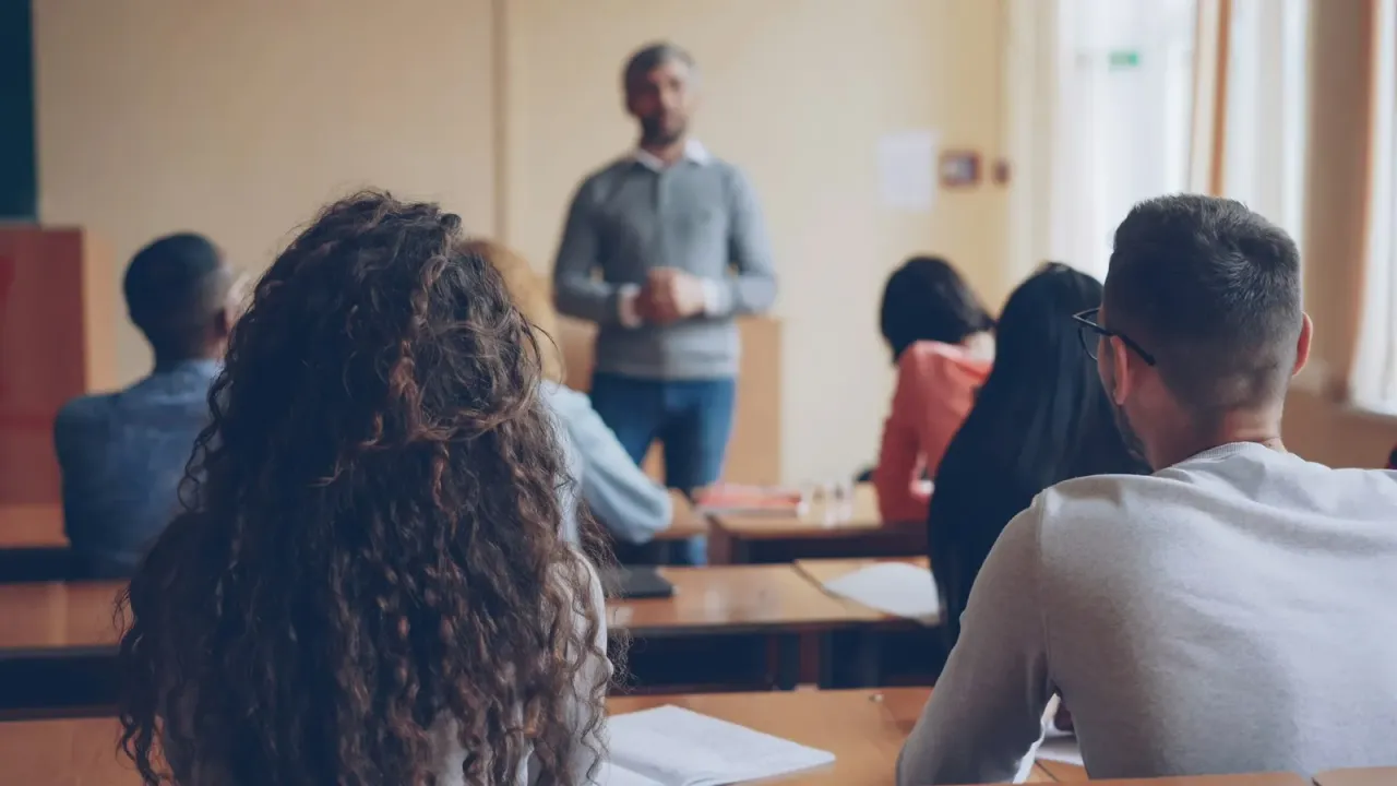 Un docente de pie al frente de un aula luminosa, conversando con la clase, que se ve desde atrás.