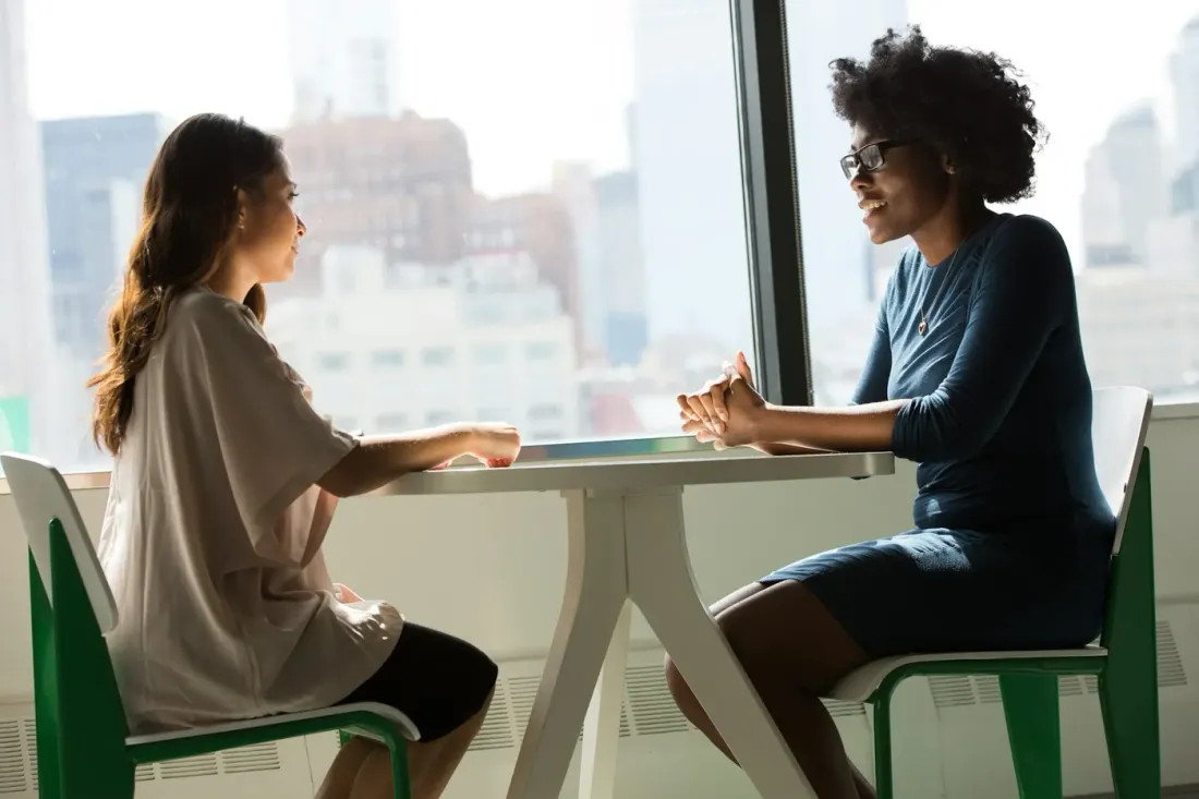 Dos mujeres conversan en una mesa junto a una ventana; una de ellas escucha.