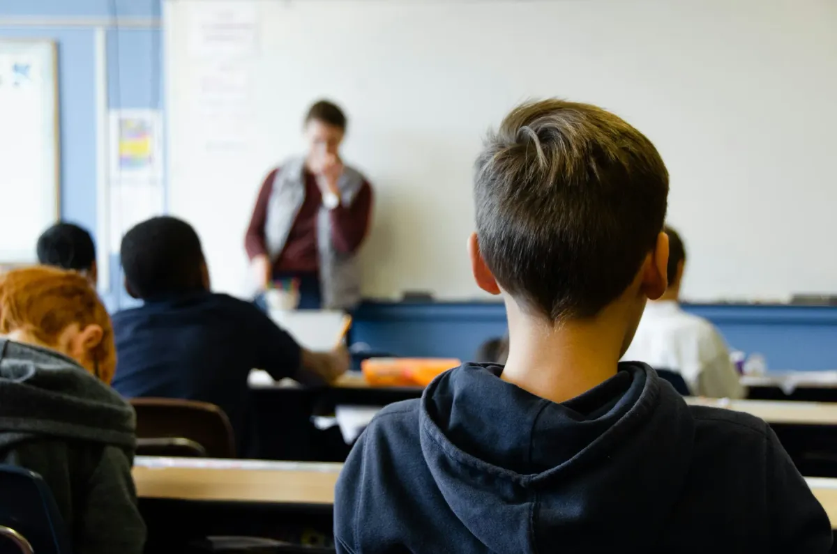 Un aula de nivel medio vista desde la última fila: un estudiante en su pupitre y, al frente, el docente fuera de foco.