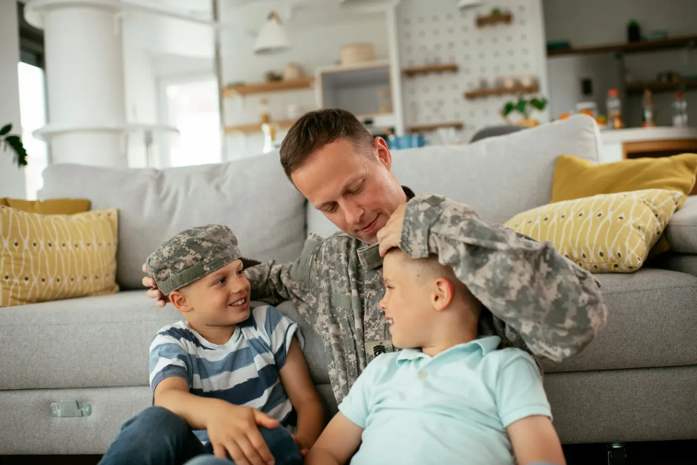 A service member in uniform sitting on the floor at home with two young boys, one of them wearing his cap.