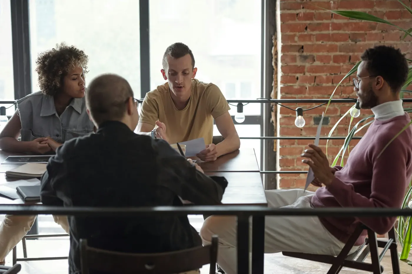 Four colleagues around a table in a bright loft office with a brick wall and big windows, mid-discussion over printed notes.