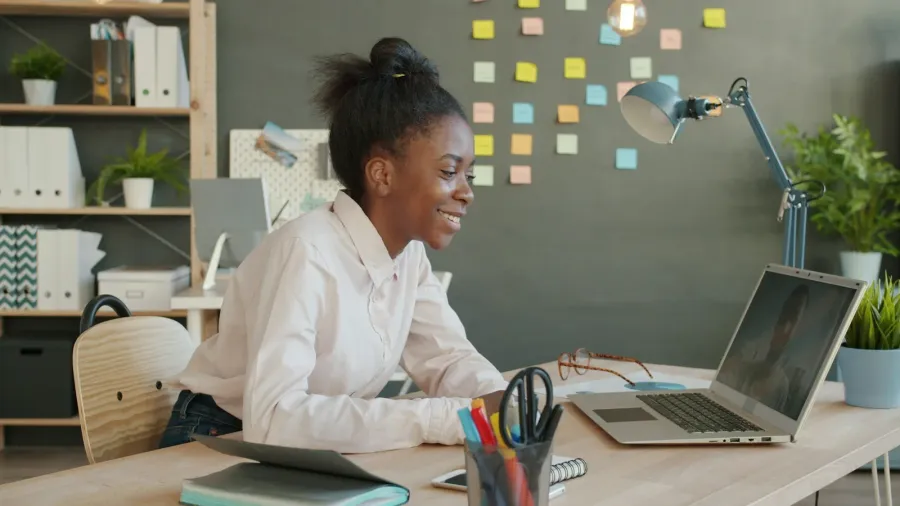Una mujer en un escritorio junto a una ventana sonríe a su laptop durante una videollamada.